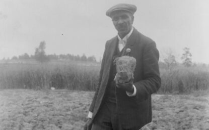 George Washington Carver standing in a field, probably at Tuskegee, holding a piece of soil, 1906.