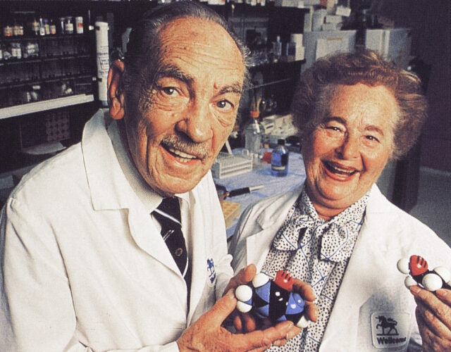 Hitchings and Elion in a lab wearing lab coats and smiling. They both hold black, blue, red, and white plastic models